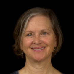 Cathleen McCarthy, a woman with chin-length light-brown-and-grey straight hair smiles at the camera against a black background