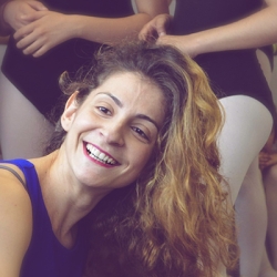 Headshot for Katerina Bousleli, she is wearing a purple dance top and smiling at the camera, her hair is loose and there are other dancers standing behind her, perhaps in a rehearsal room.